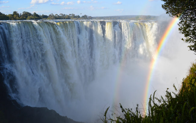 Victoriafälle in der Regenzeit mit schönem Regenbogen am rechten Rand bei blauem Himmel